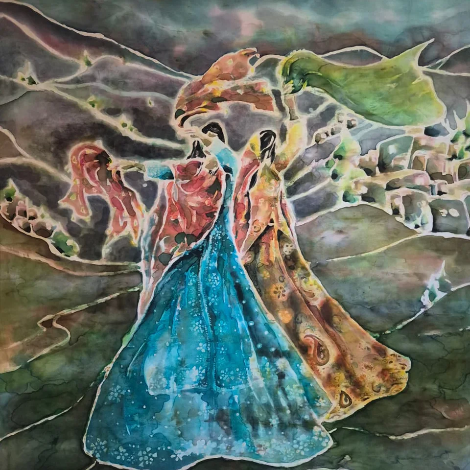 A square silk painting of two figures in traditional Lori dress, dancing close together in the wind among gray-green mountains. One wears a long turquoise dress scattered with small white flowers; the other wears a golden robe patterned with paisleys. Coral and green scarves stream from their heads, and a village of small stone houses climbs the hillside behind them. Pale resist lines outline every shape.