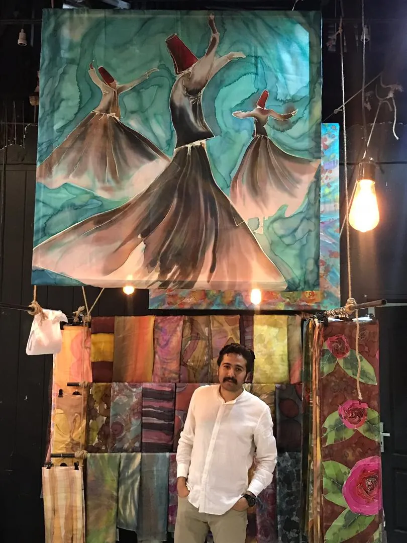 Arvin, in a white linen shirt, standing beneath a large silk painting of three whirling dervishes, with silk scarves and painted panels hanging behind him.